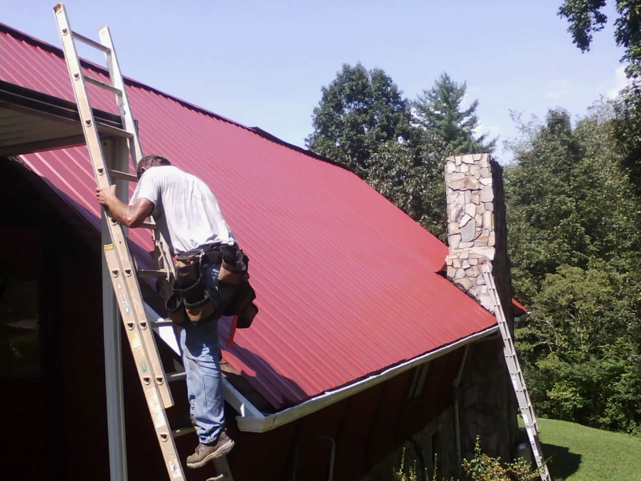 Roofer on a ladder beside a red metal roof and stone chimney — Soffit & Fascia Repair by Roofing Brothers in Nellysford